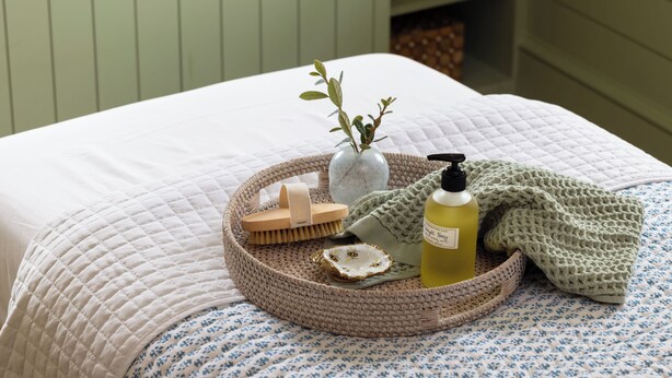 A neatly made bed with a white quilted bedspread, a woven tray with toiletries, a vase with greenery, and a crocheted blanket. - The Dunlin, Auberge Collection (Johns Island)