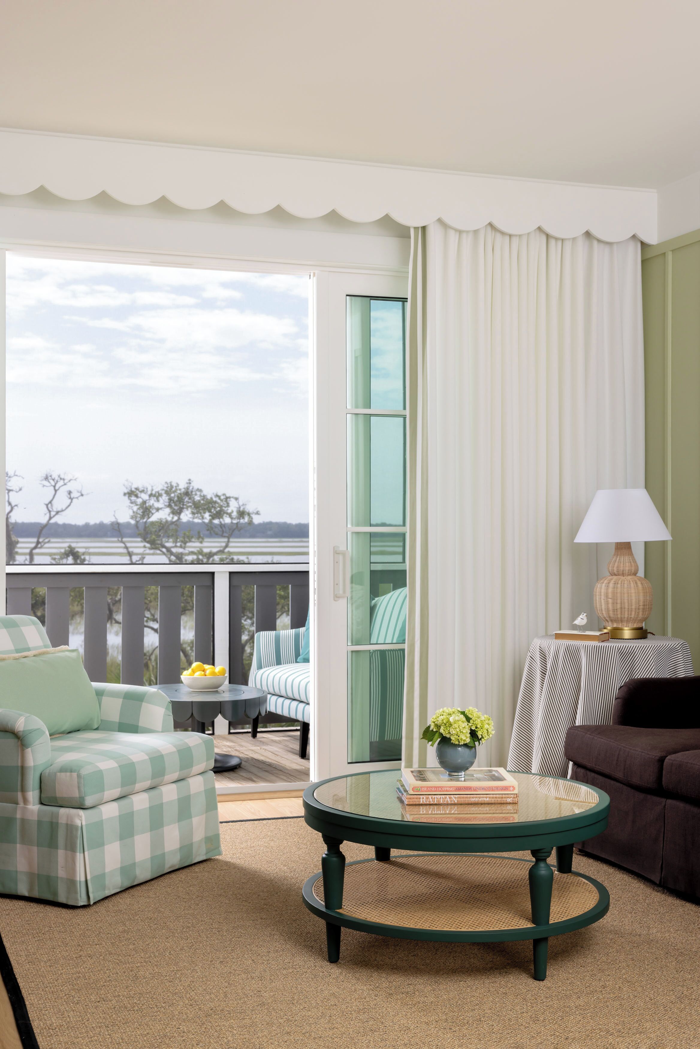 A room with a checkered armchair, a round table with a lamp, a view of the outdoors through a window, and a balcony with a railing.