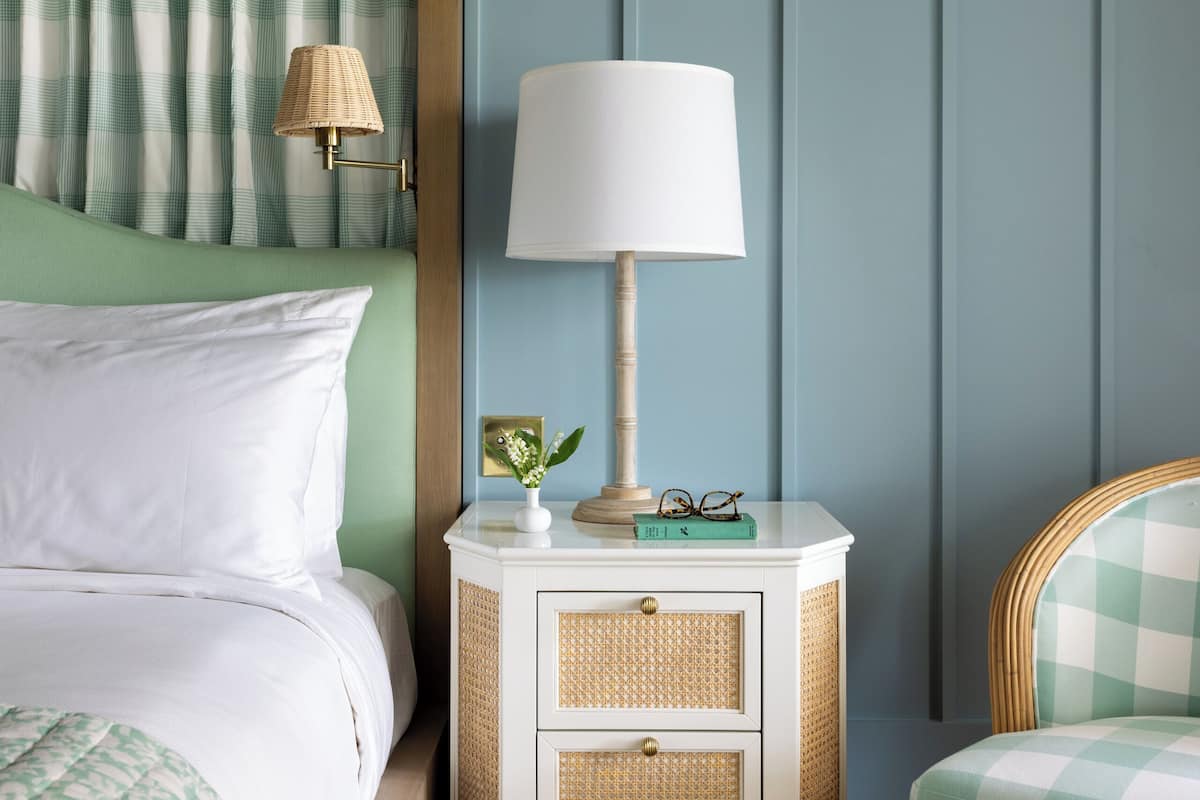 A neatly arranged bedroom with a bed, a bedside table with a lamp, a wicker chair, and a wall with vertical paneling.