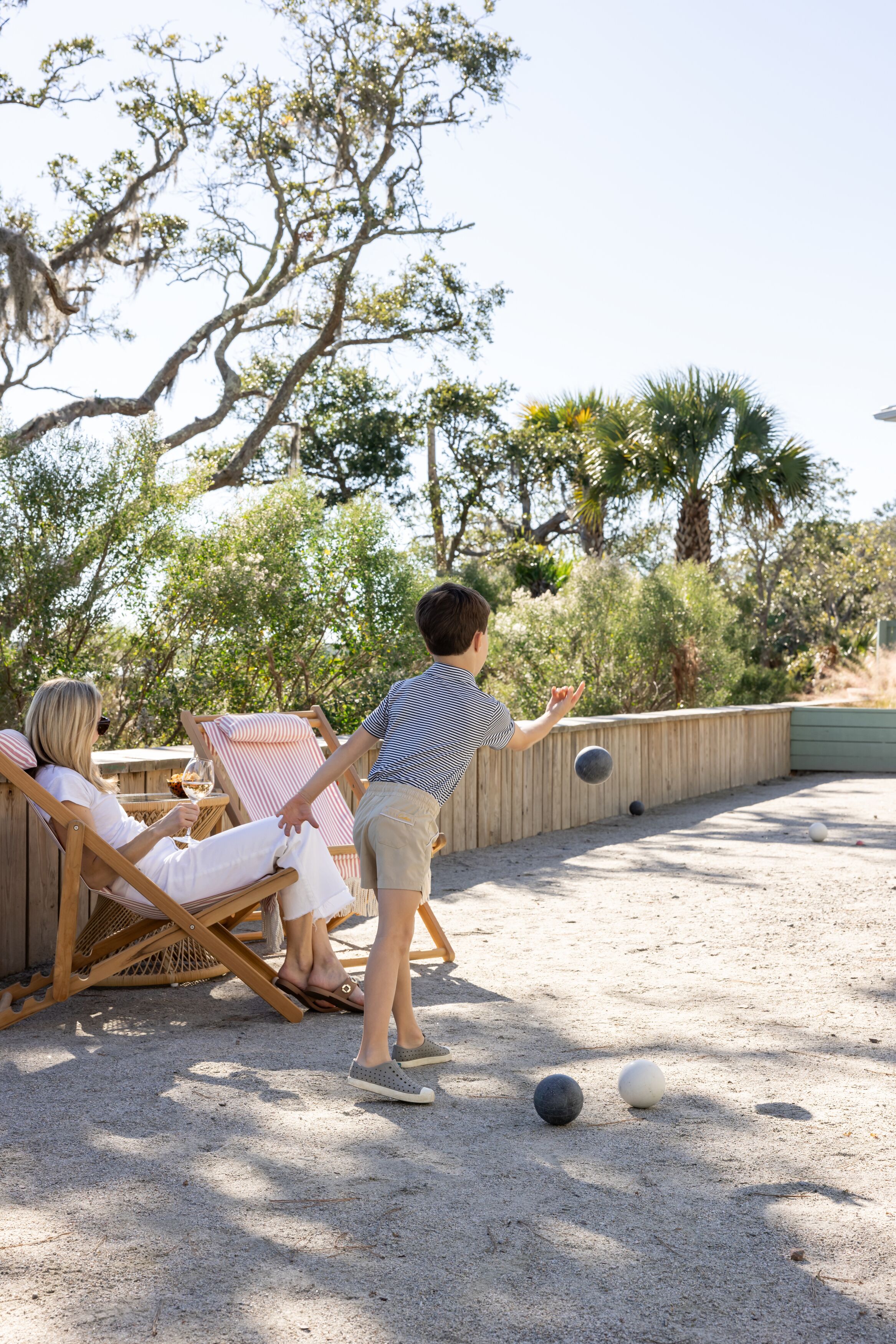Una terraza de madera con dos sillas reclinables, una mesa y una cancha de arena para croquet.