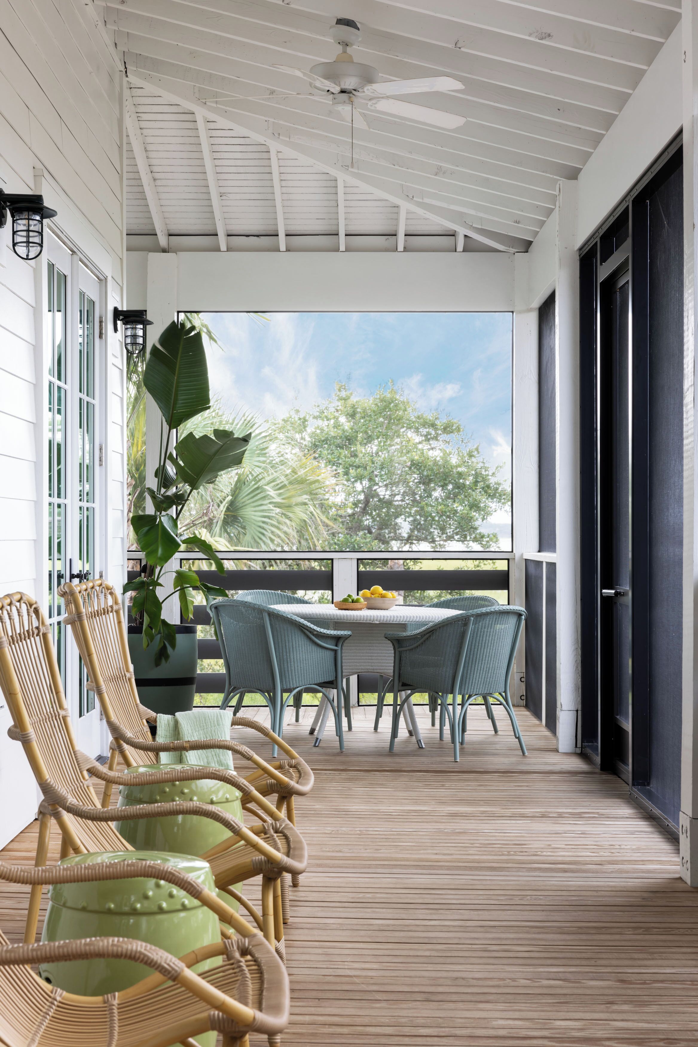 A covered porch with wicker chairs and a table, a potted plant, and a view of trees and sky.