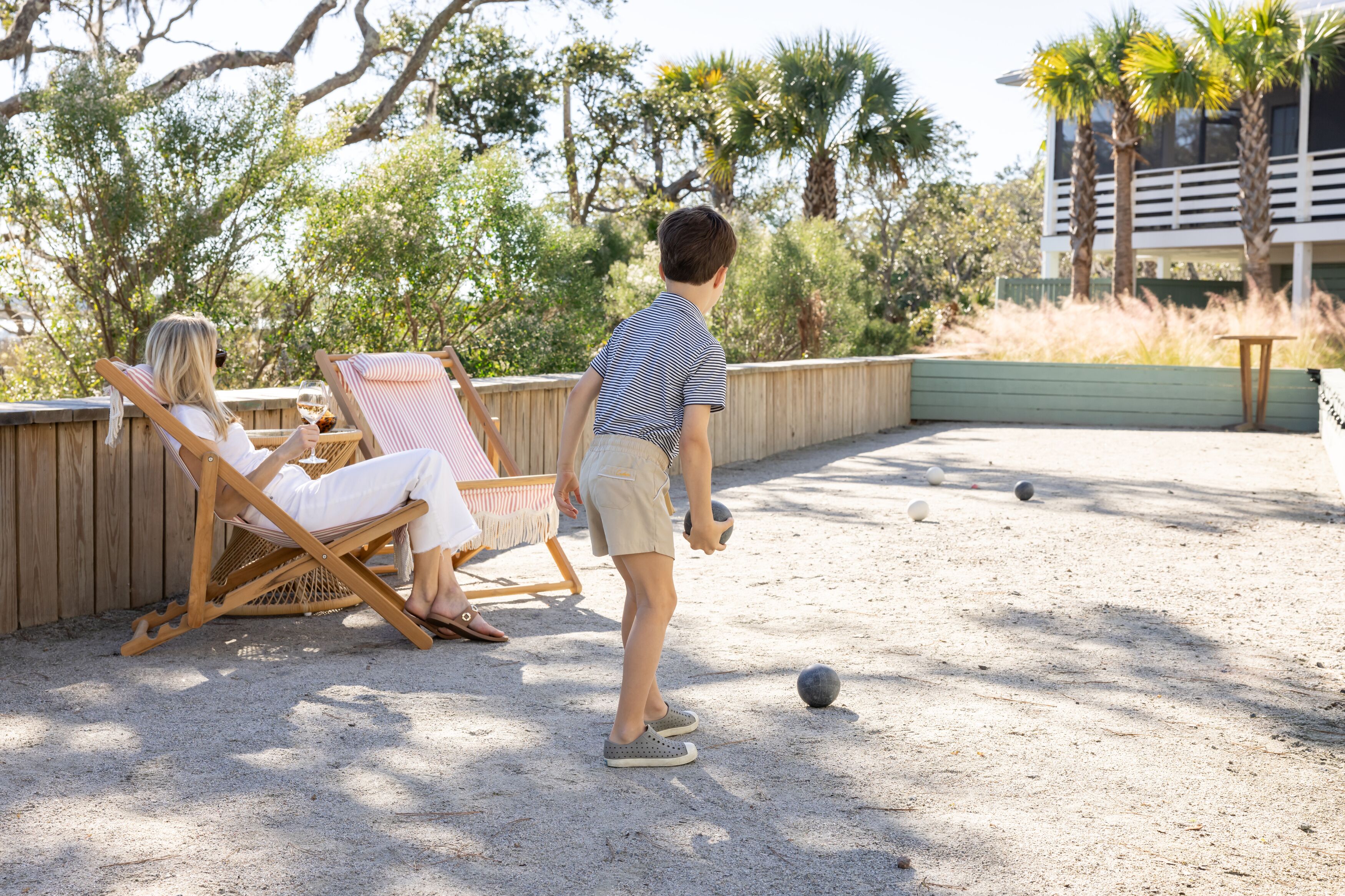 A person playing croquet on a sandy court with a wooden fence and palm trees in the background.