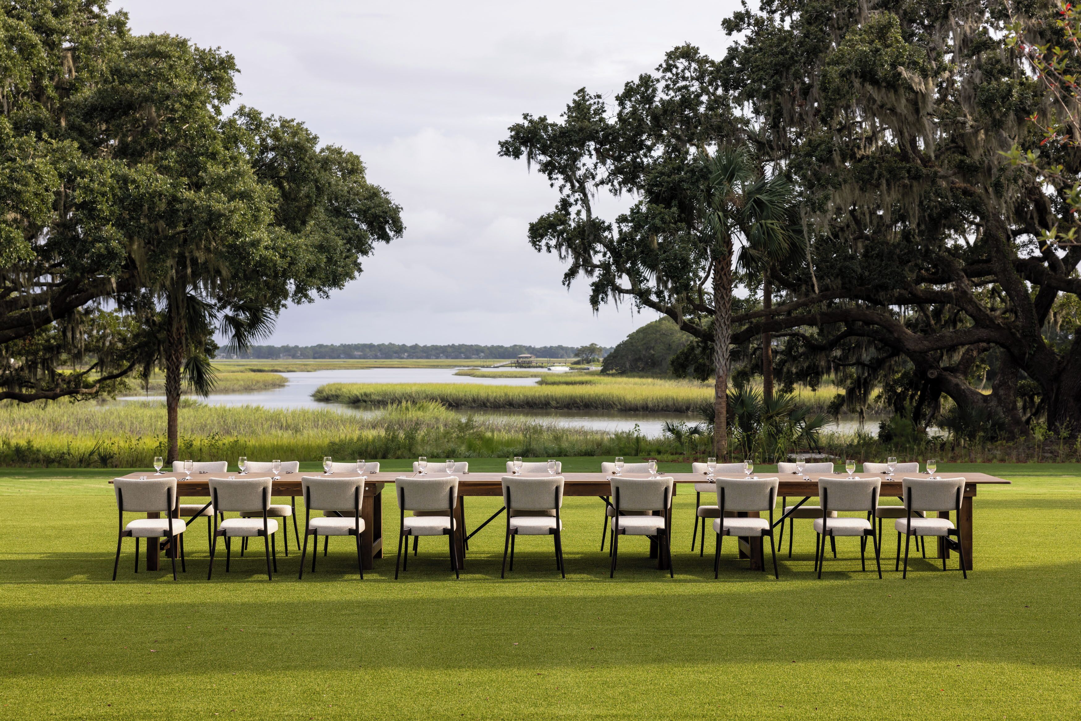 A dining table set up outdoors with chairs, surrounded by trees and a view of a water body.