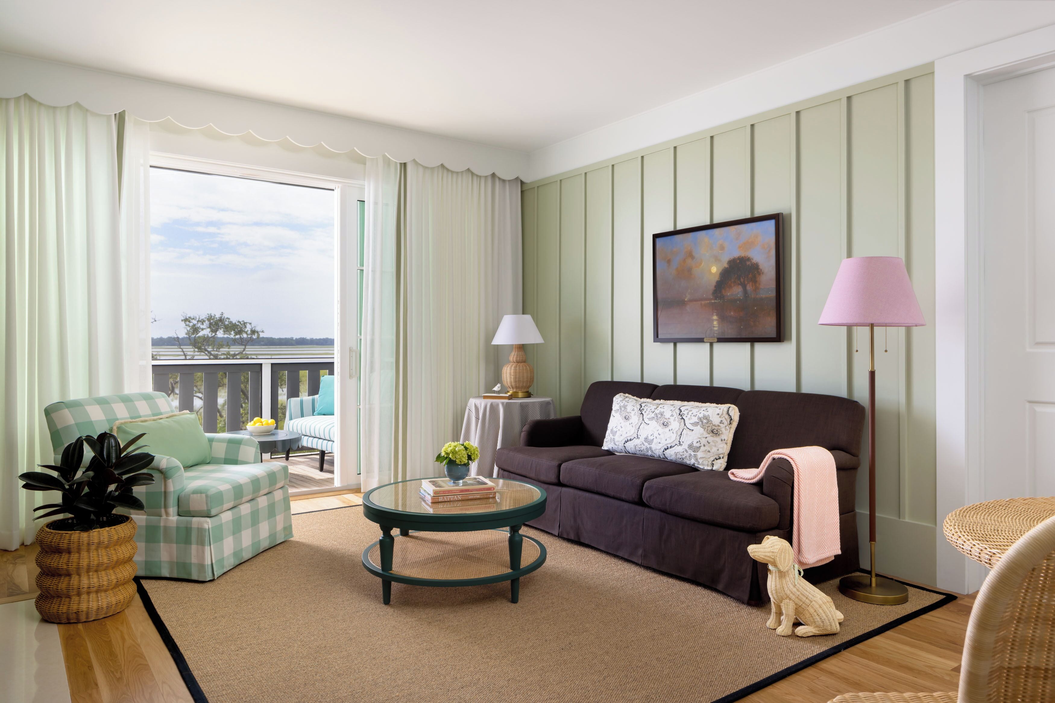 Un salon avec un fauteuil à motifs à carreaux, une table basse ronde, un canapé foncé, une petite table avec une lampe et un tableau encadré au mur.