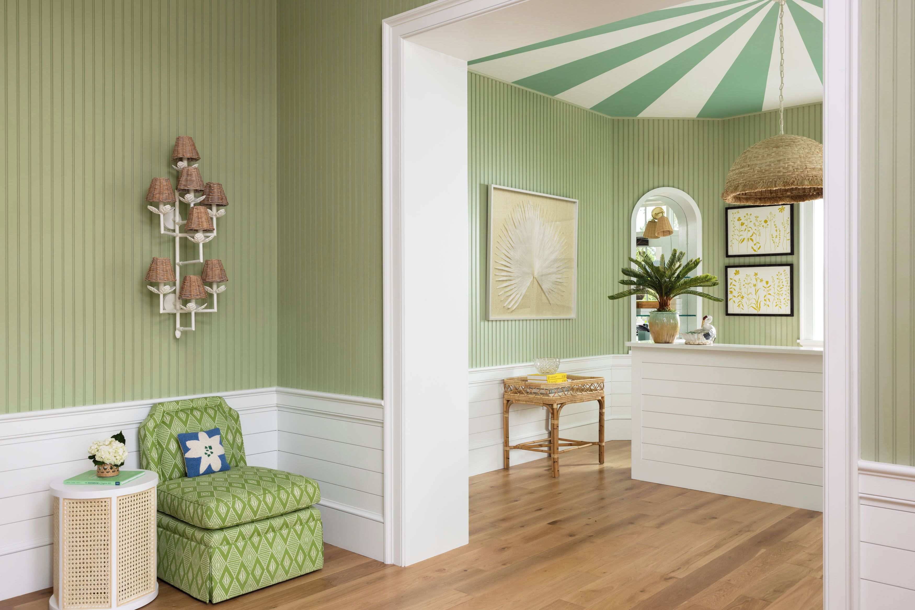 Een kamer met een kleurenspel van groen en wit, met een patroonstoel in het groen, een rieten bijzettafeltje en een hanglamp van riet.