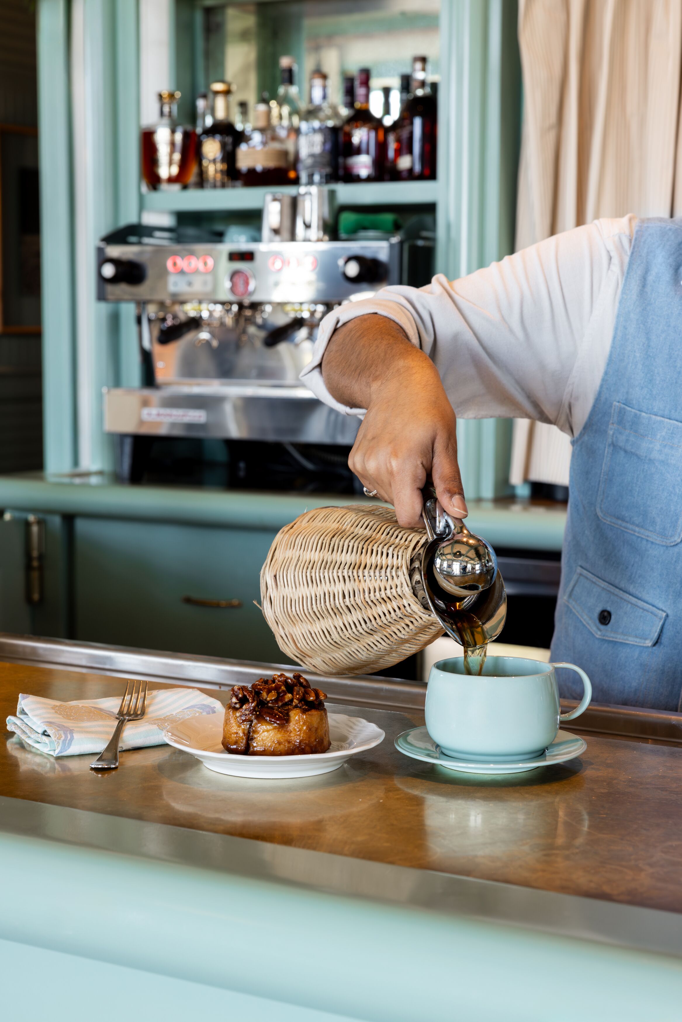 A person pouring a beverage into a cup on a table with a pastry and a fork.
