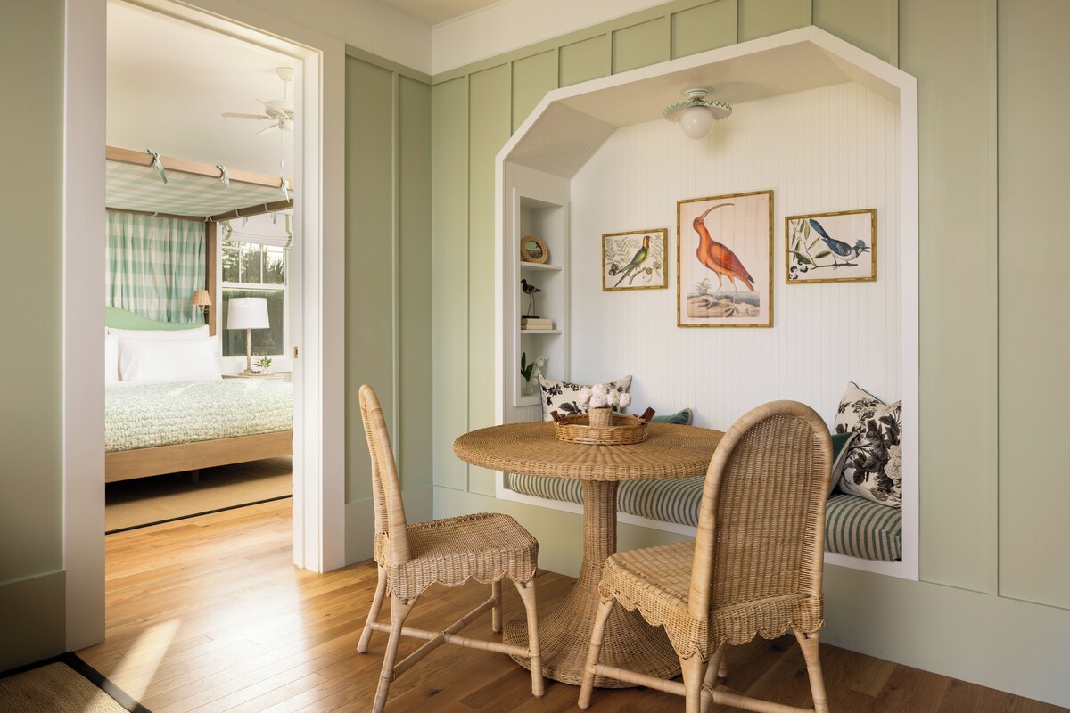 Una habitación con una mesa redonda y sillas de mimbre, una cama con colcha verde a cuadros, y estantes en la pared con obras de arte enmarcadas.