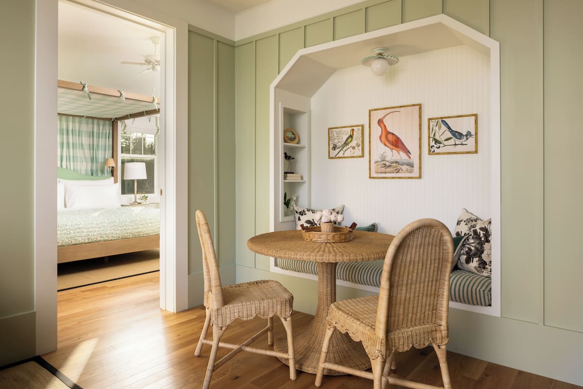 A room with a round table and wicker chairs, a bed with a green checkered bedspread, and wall-mounted shelves with framed artwork.
