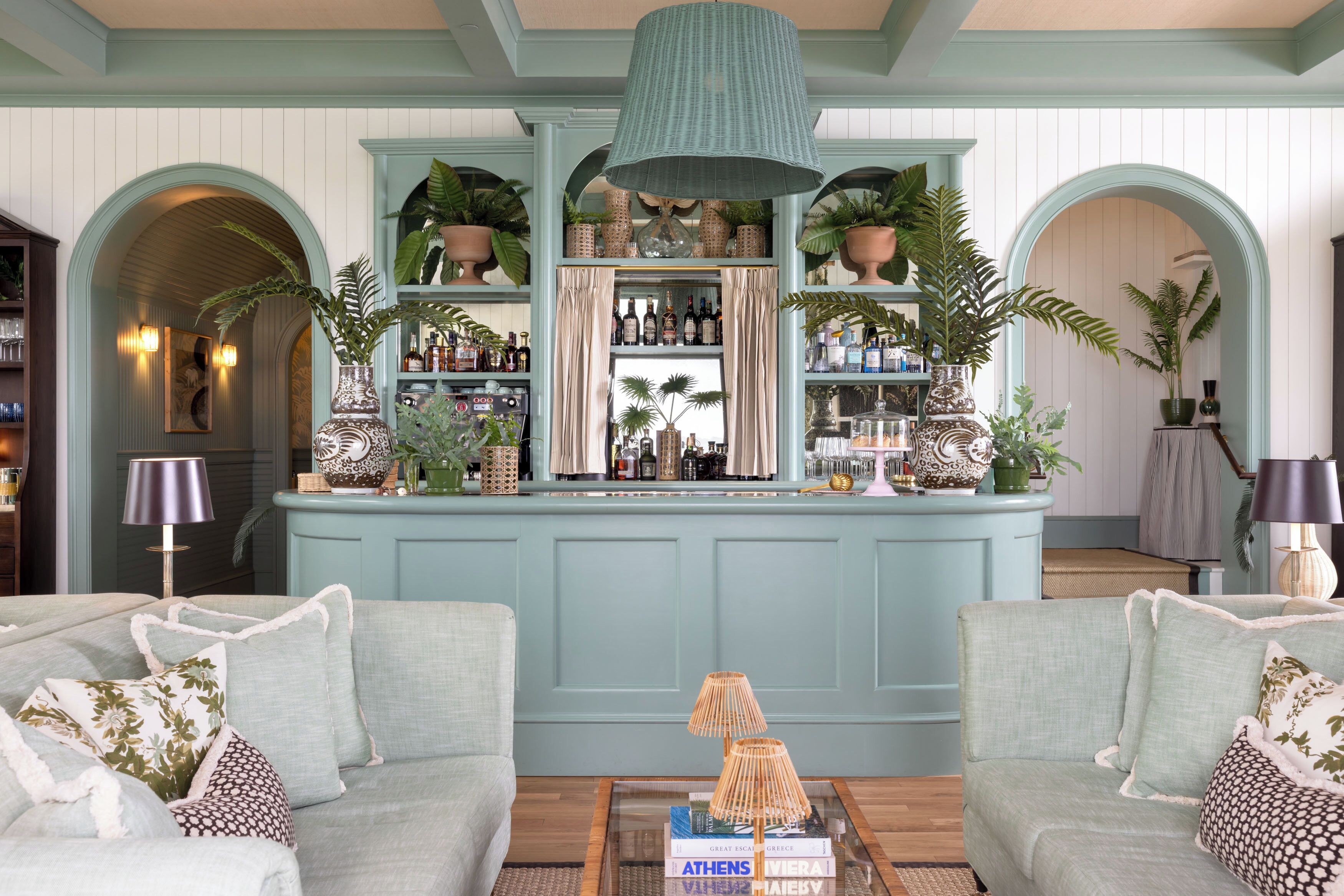 Un espace bar avec un fond bleu clair, des étagères remplies de bouteilles et des plantes en pot. On y trouve deux canapés ornés de coussins à motifs.