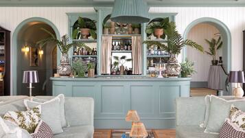 A bar area with a light blue backdrop, shelves with bottles, and potted plants. There are two sofas with patterned cushions in front.
