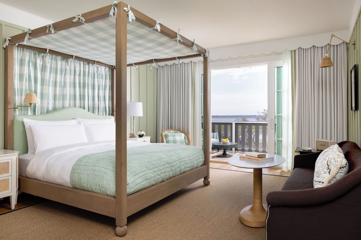 A four-poster bed with a checkered canopy, a wooden nightstand with a lamp, a round coffee table, a sofa, and a view of the sea through large windows.
