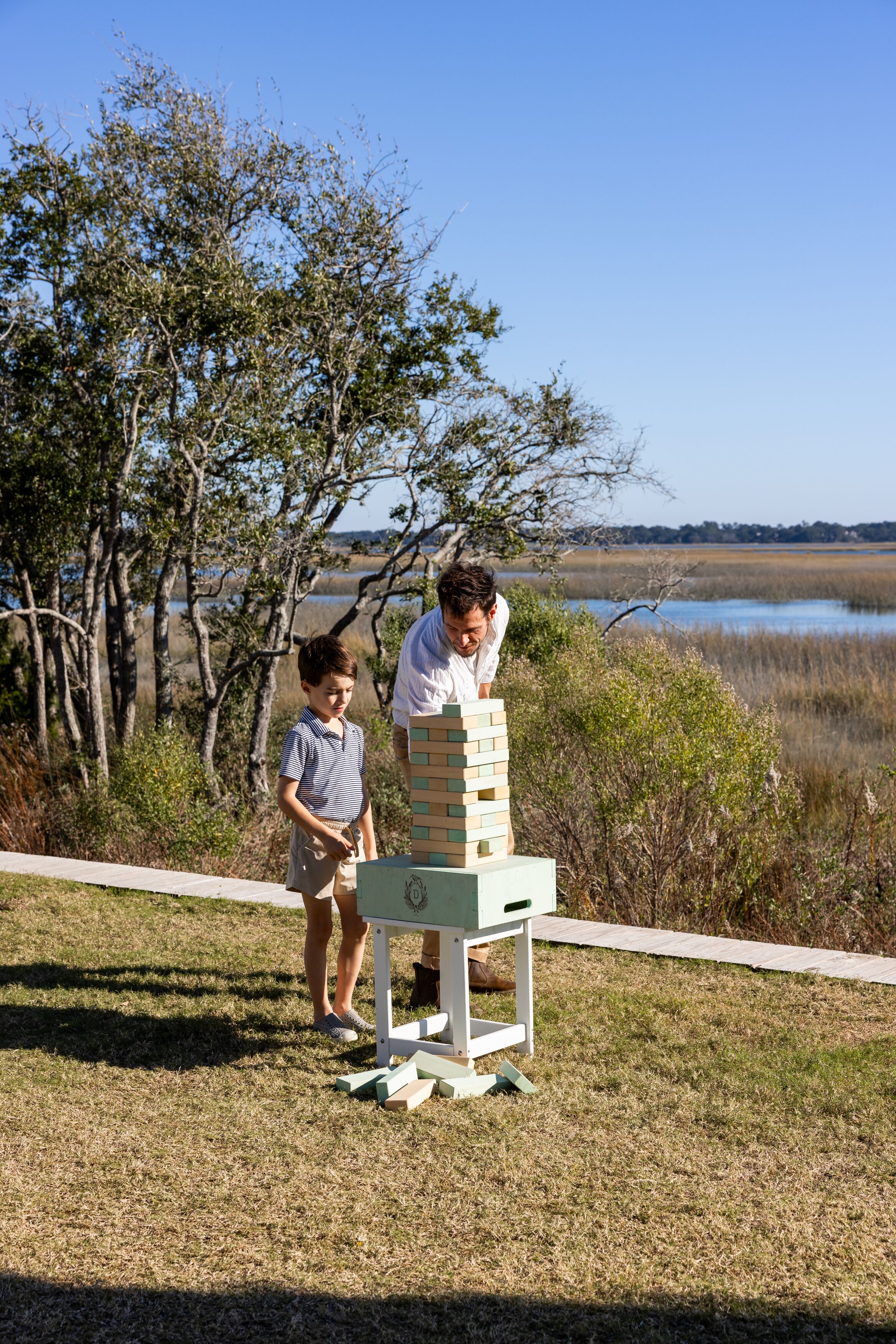 Twee personen spelen Jenga in een open setting, met een waterpartij en bomen op de achtergrond.