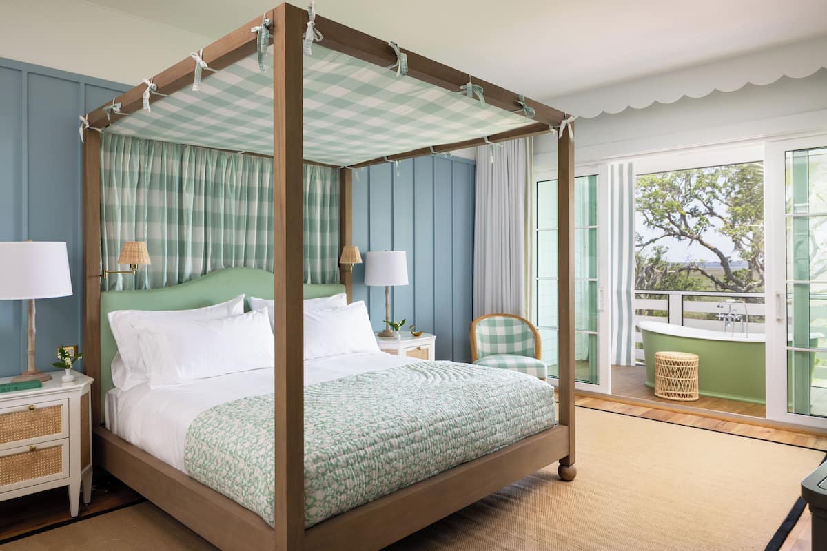 A four-poster bed with a checkered canopy, a nightstand with lamps, a wicker chair, and a view of the outdoors through a balcony.
