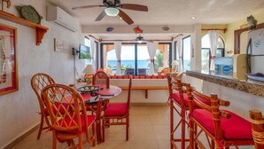 A dining area with a view of the ocean, wooden chairs with red cushions, a ceiling fan, and a kitchen counter with a refrigerator. - Relax in Your Private Beachfront Retreat, Surrounded by Breathtaking Ocean Views (Akumal)