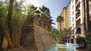 Zona de piscina con pared de roca, palmeras y un edificio con balcones.