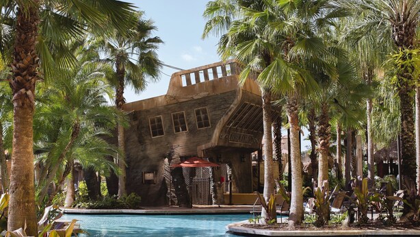 Un área de piscina con palmeras, un edificio de fachada de piedra y una estructura con techo rojo.