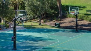 An outdoor basketball court with two hoops, surrounded by trees and a grassy area.