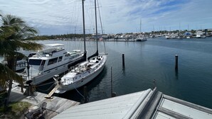 A marina with several sailboats docked, a wooden deck with outdoor seating, and a clear view of the water and distant shoreline. - Living Life Dockside (Naval Air Station Key West)