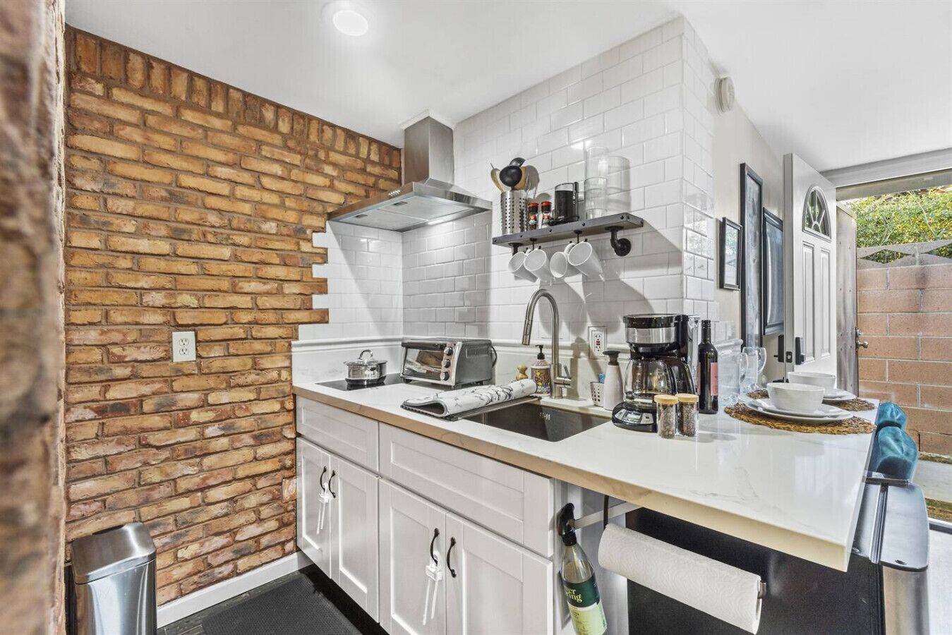 A kitchen with white cabinets, a brick accent wall, a stainless steel range hood, and a window with a view of greenery.
