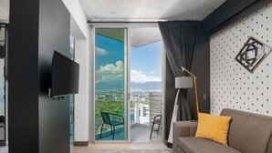 A modern living room with a grey sofa, a flat-screen TV, a floor lamp, and a balcony with a city view.