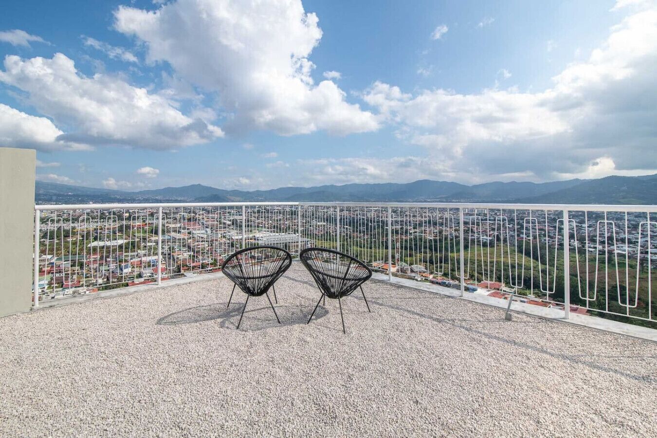 A terrace with two black chairs, a white railing, and a view of a cityscape and mountains.