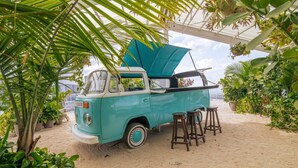 A vintage turquoise van parked in a sandy area with wooden stools, surrounded by lush greenery and potted plants.