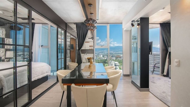 A modern hotel room with a large dining table, white chairs, a view of the city, and a balcony with a railing.