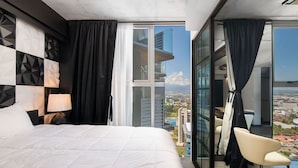 A modern hotel room with a large bed, a chair, and a view of the city through floor-to-ceiling windows.