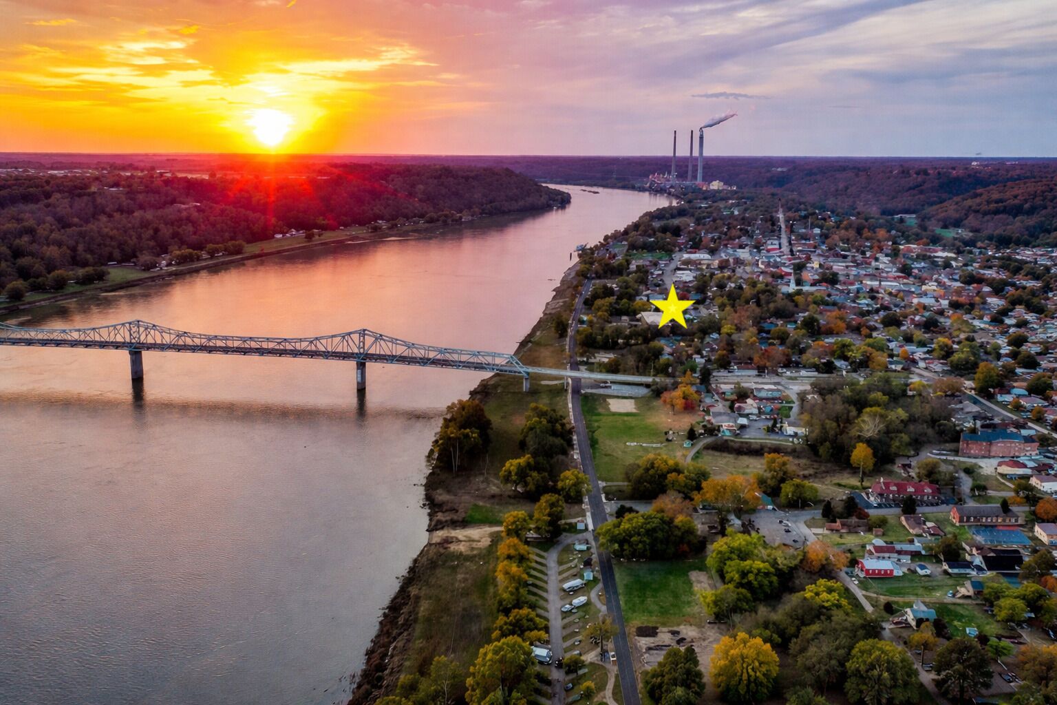 Luchtbeeld van een stad met een rivier, een brug en industriële schoorstenen op de achtergrond.