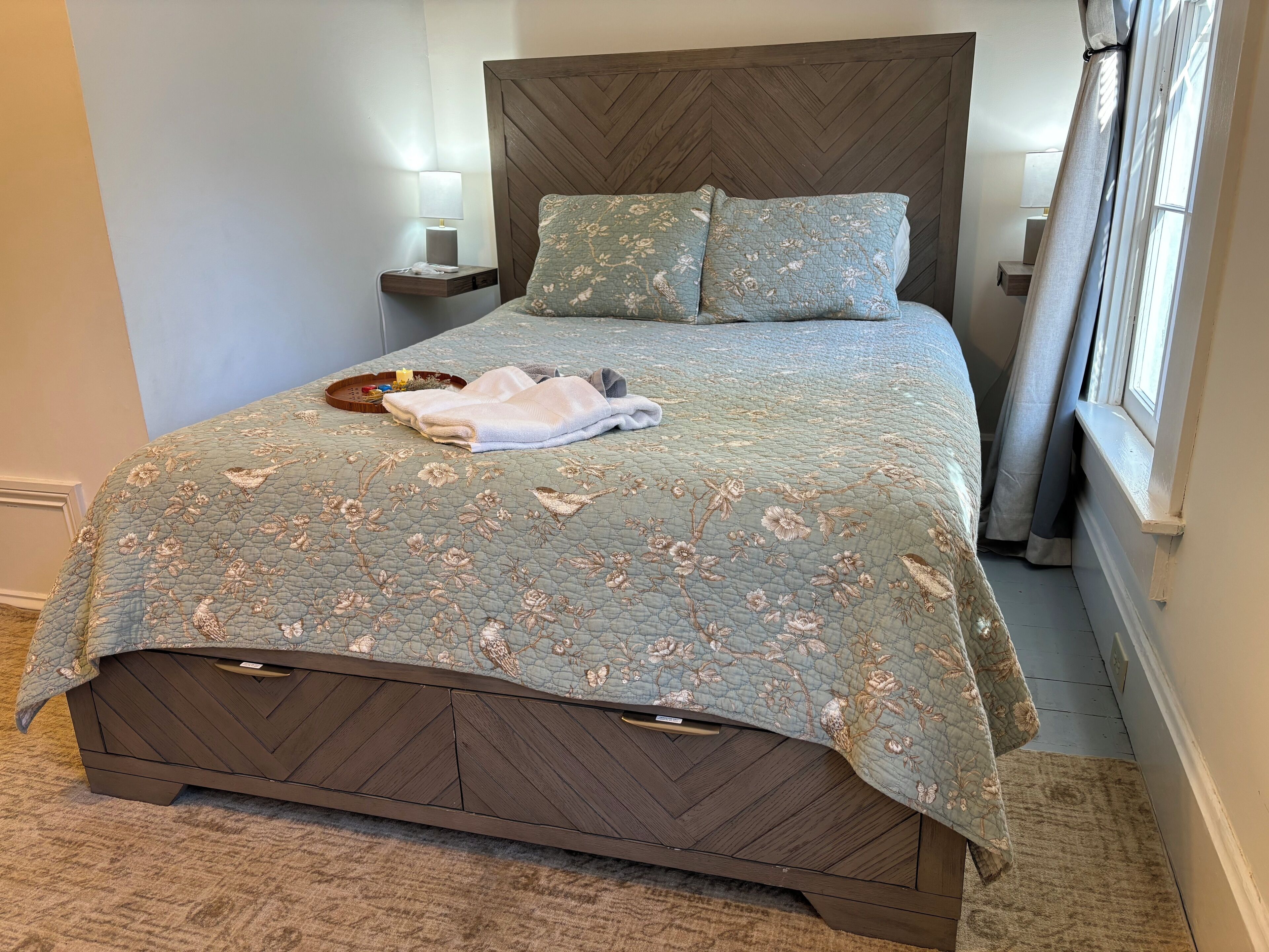 A neatly made bed with a floral quilt, a bedside table with a lamp, and a window with curtains.