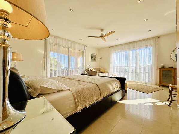 Un dormitorio amplio con una cama grande, un ventilador de techo y vistas al exterior a través de amplias ventanas.