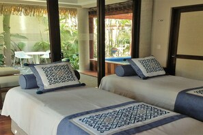 A hotel room with two beds, a small table, and a view of a tropical garden.