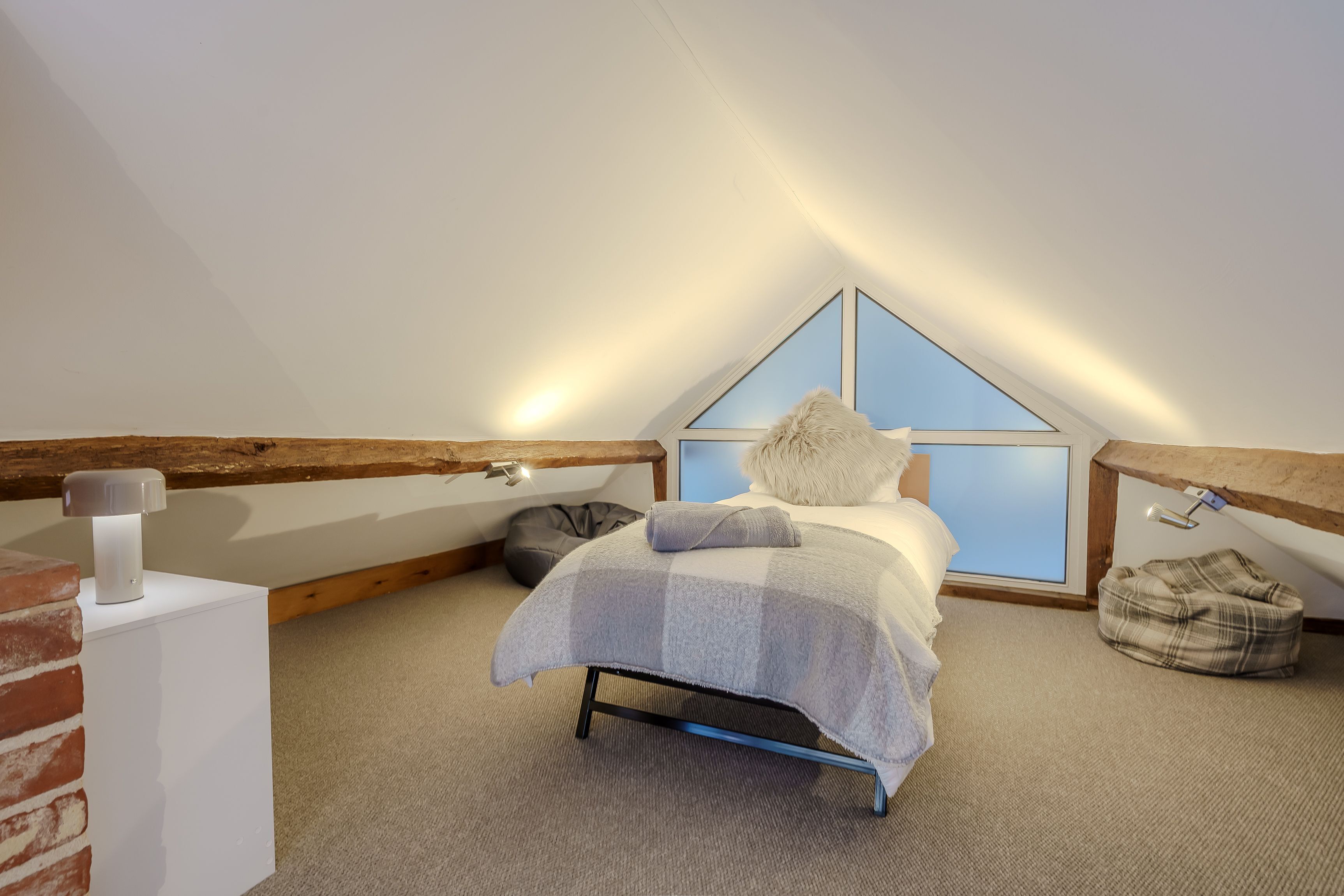 A cozy attic bedroom with a sloped ceiling, a bed with a white and gray checkered blanket, a fluffy pillow, and a small bedside lamp.