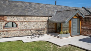 Een gebouw van steen met een schuin dak, een klein terras met een tafel en stoelen, en een deur met een raam.