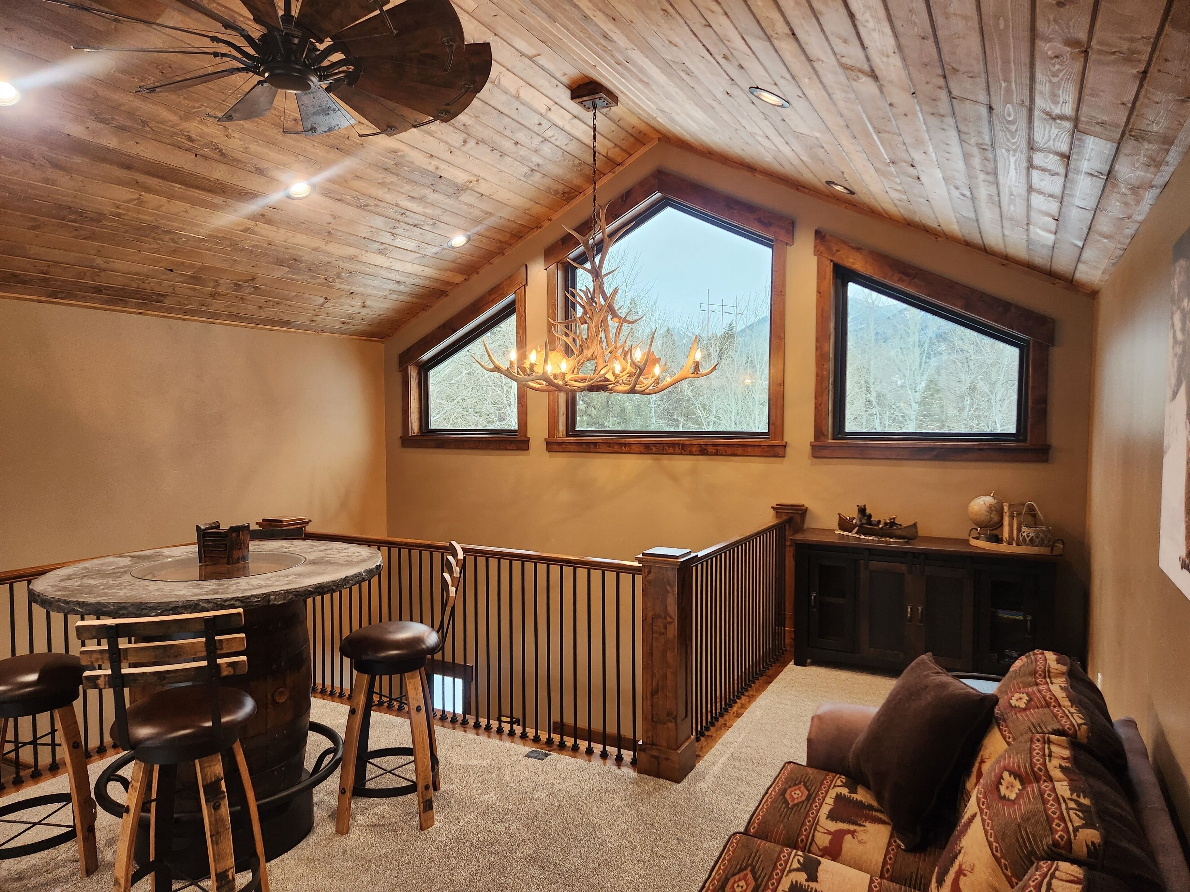 A spacious room with a wooden ceiling, a round bar area with stools, a chandelier, and large windows offering a view of trees.
