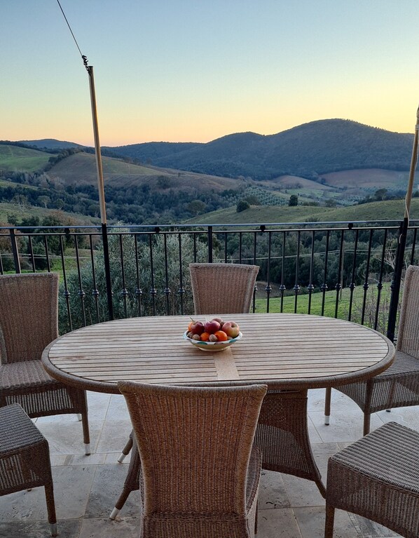 Une table ronde en bois, entourée de chaises en osier, un saladier rempli de fruits et une vue sur des collines ondoyantes.