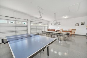 A ping pong table in a room with a dining table and chairs, white walls, and a garage door.
