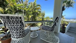 Ein Balkon mit weißen Korbstühlen und einem kleinen Tisch, umgeben von tropischen Pflanzen und mit Blick auf eine schöne Aussicht.