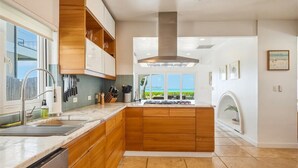 Una cocina moderna con armarios de madera, un extractor de acero inoxidable, un ventanal con vistas al océano y una isla integrada con un lavabo.
