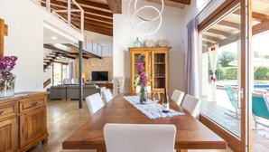 Un vaste espace dédié à la salle à manger, avec une table en bois, des chaises blanches et une vue sur la piscine, visible à travers de grandes portes-fenêtres coulissantes.