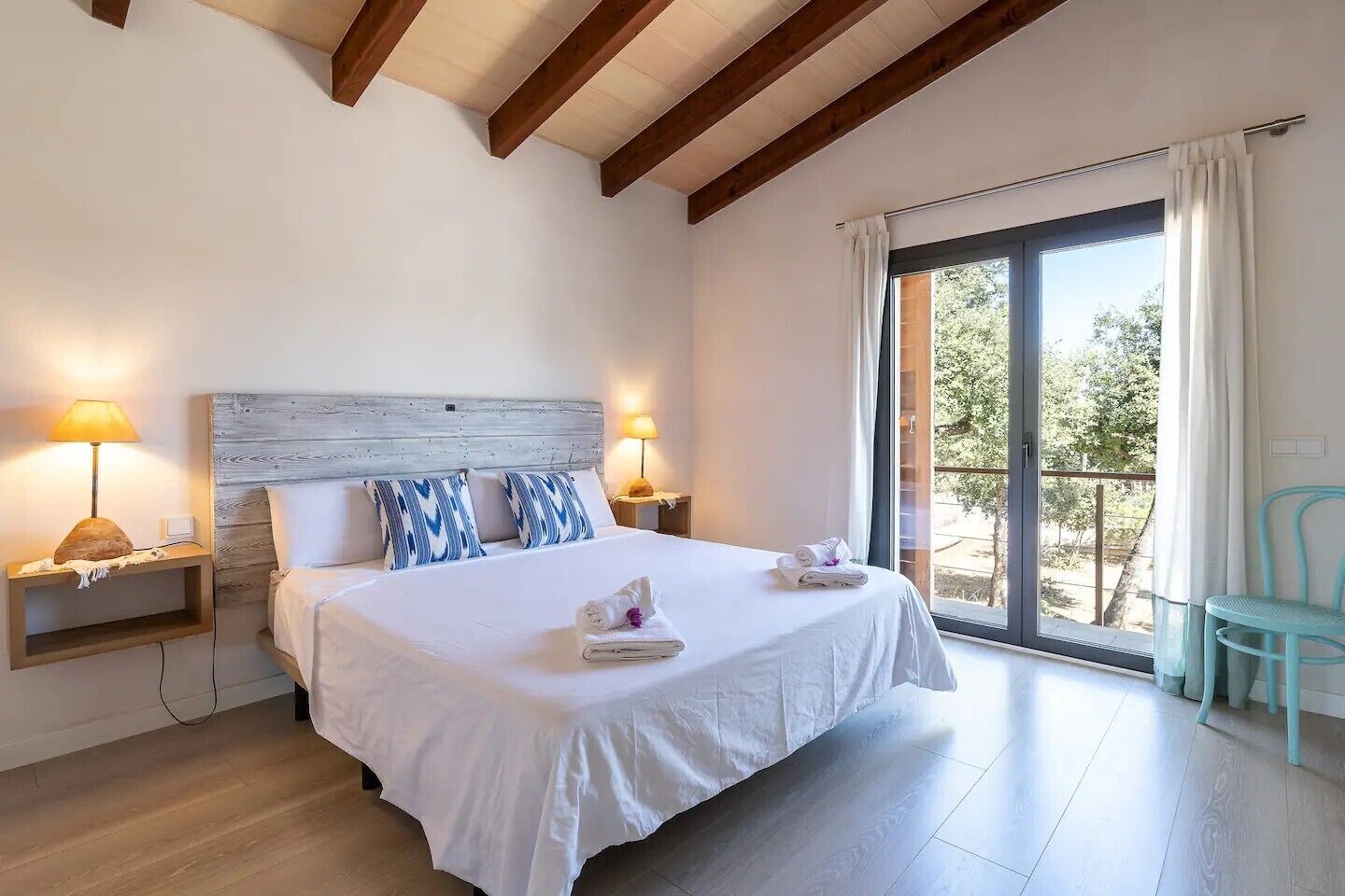 Un dormitorio con una cama grande, dos mesitas de noche con lámparas, vistas a través de un ventanal corredero y un balcón con una silla.