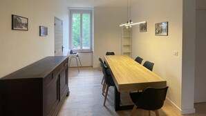 Ein Essbereich mit einem langen Holztisch, schwarzen Stühlen, einem weißen Stuhl, einem kleinen weißen Regal und einem Fenster mit Blick ins Grüne.