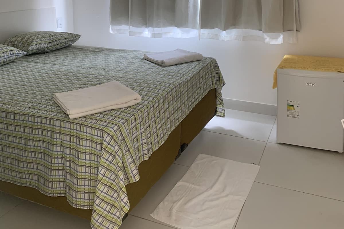 Een slaapkamer met een bed, kussens, een nachtkastje, een raam met gordijnen en een radiator.