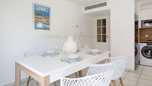 A dining area with a white table set for four, white chairs, a poster of Cannes, and a laundry area with a washing machine. - Palazzio Re-2753-Modern apartment close to the Port (Cannes)