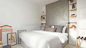 Un dormitorio con una cama, un perchero de madera, una mesita y un estante en la pared con adornos.