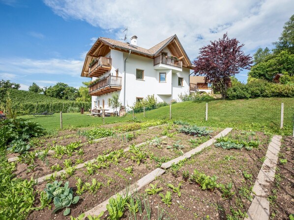 A two-story house with a balcony, surrounded by a garden with vegetable plots.