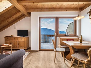 A cozy room with a wooden ceiling, a television, a dining table with chairs, and a view of mountains through large windows.