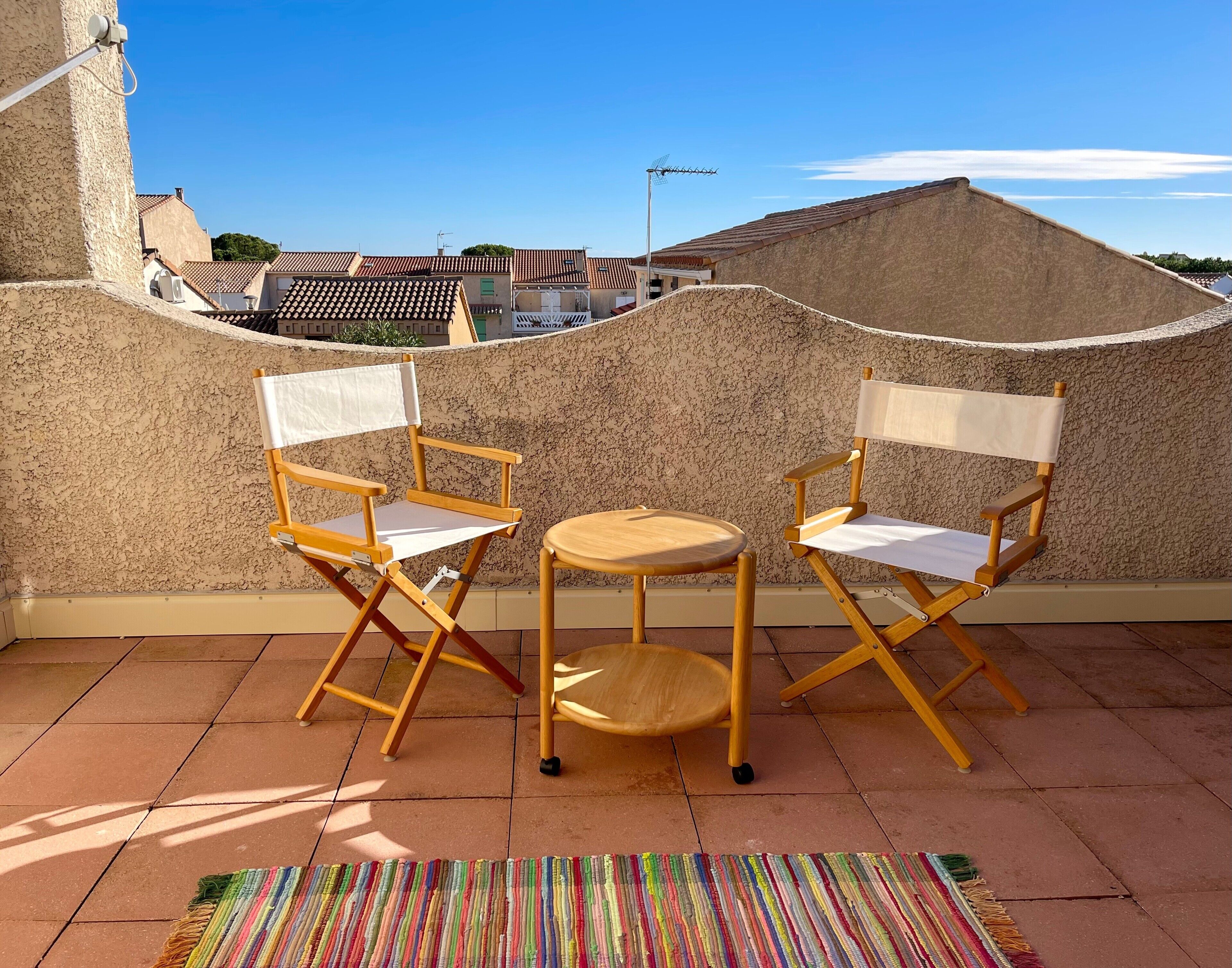 A terrace with two wooden director's chairs and a small round table, a colorful striped rug, and a view of buildings in the background.