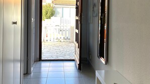 A hallway with a tiled floor, a white bench with a folded blanket, and a door leading to an outdoor area with a white fence and buildings in the background.