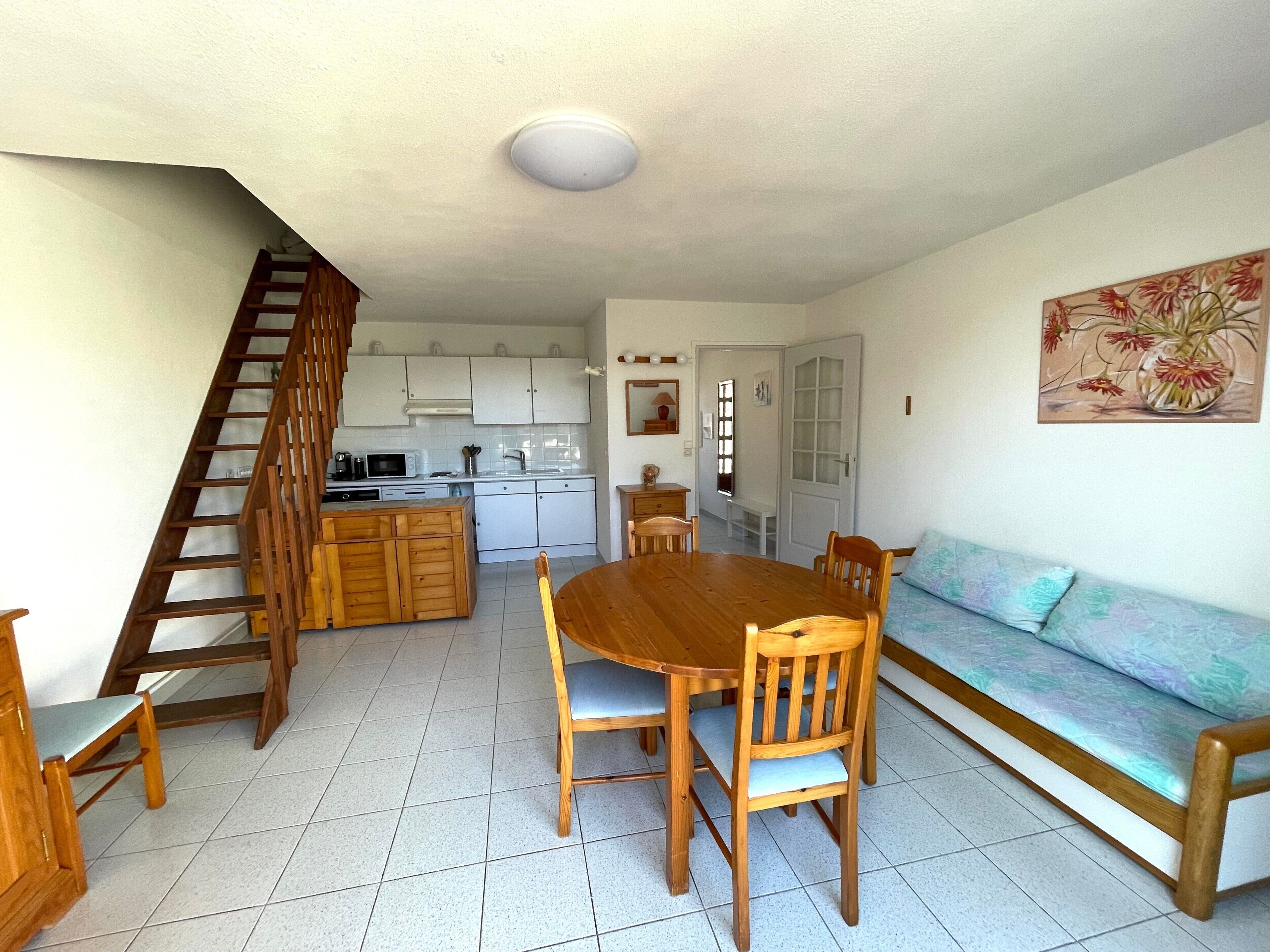 Une cuisine avec des meubles blancs, une table à manger en bois et des chaises, un escalier et un coin salon avec un canapé.